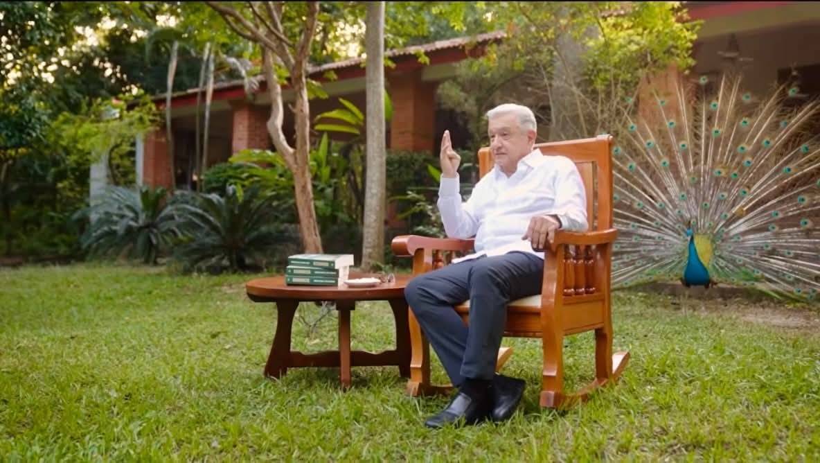 Andres Manuel López Obrador sentado en una silla de madera a su espalda un pavo real abriendo su plumaje